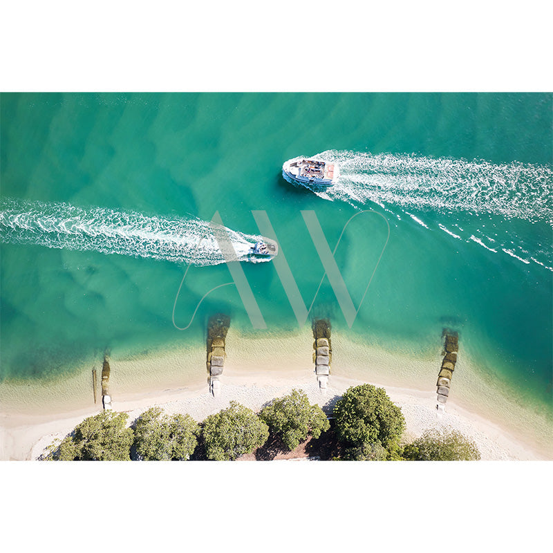 Aerial view of two boats creating wakes on turquoise water near a sandy beach with lush green trees. Remnants of old wooden structures are partially submerged in the water.