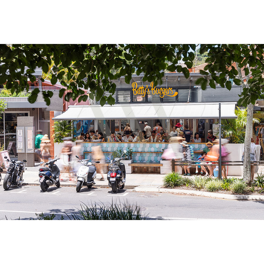 A lively street scene in front of a burger restaurant with a teal and white tiled facade. People are seated, eating, or walking by, and several scooters are parked along the sidewalk. Lush greenery frames the view.