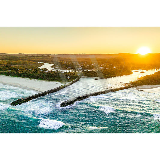 Aerial view captured in Brunswick Heads Drone Image 0240 by AWP Image Library shows a coastal landscape at sunset with two rock breakwaters in the ocean, waves gently lapping, and a golden glow from the setting sun over the water and lush green scenery.