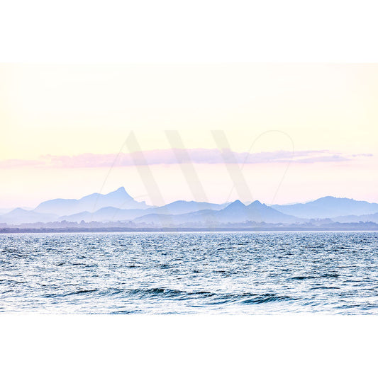 The Byron Bay Image 7104 by AWP Image Library captures a serene ocean view at Wategos Beach with gentle waves, distant hazy blue mountains, and a pastel sunset sky.