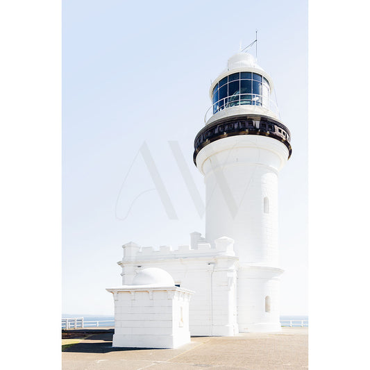 The Byron Bay Lighthouse Image 8457 from the AWP Image Library shows a sunlit white lighthouse with a black band at the top, standing against a clear blue sky and casting shadows on the ground in a coastal scene without any visible ocean.