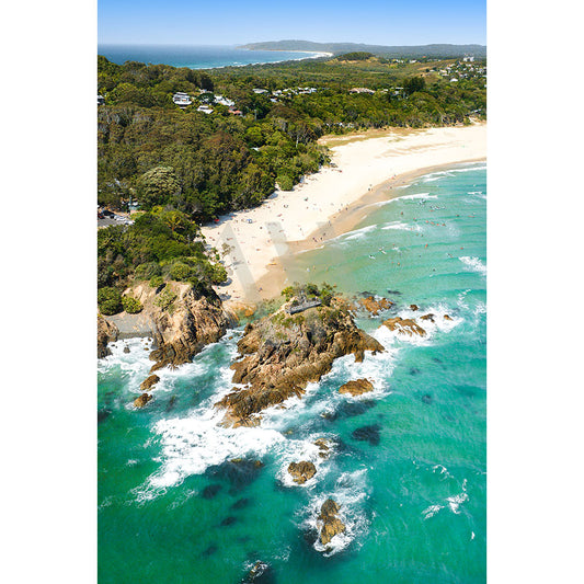 The Byron Bay The Pass Drone Image 0519 by AWP Image Library showcases a stunning aerial view of a scenic coastline with a sandy beach, lush green trees, and turquoise ocean waves crashing against rocky outcrops as people enjoy the sunny day.