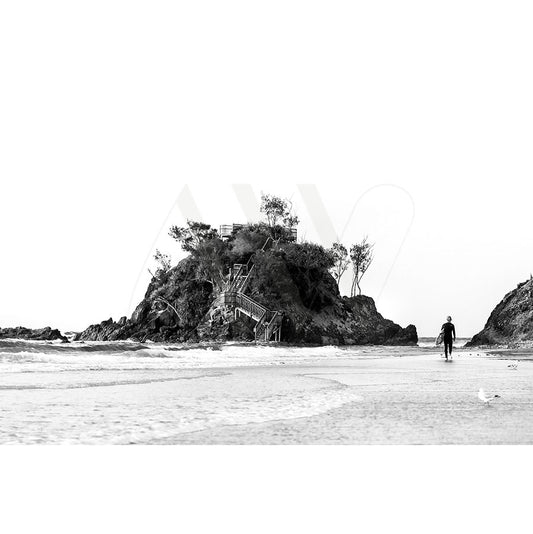 The AWP Image Library presents the Byron Bay The Pass B&W Image, depicting a small rocky island with trees, a staircase leading up, and a person walking along the sandy beach towards the island while a seagull stands near the shoreline.