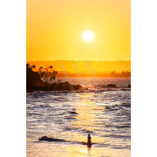 Byron Bay Wategos Image 6982, from AWP Image Library, captures a person standing in the ocean at sunset with an orange sky. Silhouetted rocks and trees create a stunning backdrop as waves gently lap the shore.