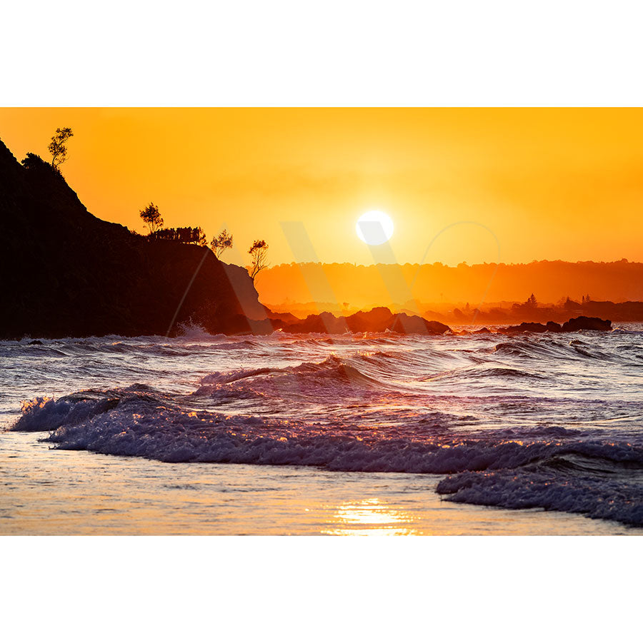 The Byron Bay Wategos Beach Image 7073 by AWP Image Library captures a sunset over a serene beach, with waves lapping gently against the shore, a vibrant orange sky, and silhouetted trees on the rocky coastline.