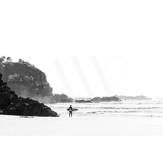 The Byron Bay Whites Beach Image 5663 by AWP Image Library features a black and white scene of a surfer with a surfboard heading to the ocean, rocky cliffs on the left, gentle waves, sparse trees on the cliff, creating a serene and expansive atmosphere.