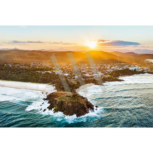 The Cabarita Beach Drone Image 0432 by AWP Image Library showcases a coastal town at sunset, with the sun setting over distant hills, illuminating the ocean. Waves crash against a rocky promontory as palm tree shadows dance on the sand, and houses nestle among lush greenery.