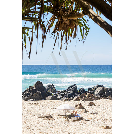 Cabarita Beach Image 4869 by AWP Image Library captures a serene beach scene with a person relaxing under a small white umbrella on sandy shores. Ocean waves gently crash against black rocks, while leafy branches of a tree frame the image against a clear blue sky.