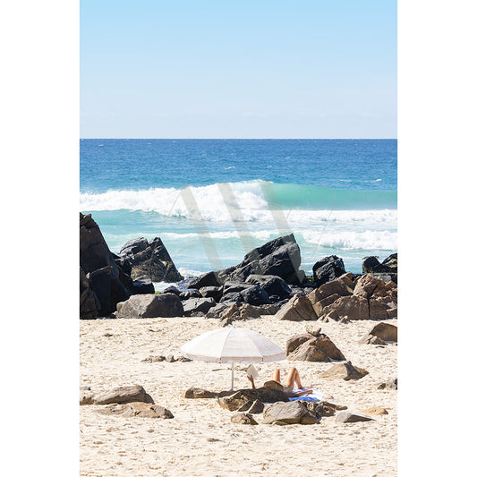 Cabarita Beach Image 4874 by AWP Image Library shows a person relaxing under a white umbrella on sandy shores, lying on a towel near large rocks. Waves crash in the background against the backdrop of a clear blue sky.