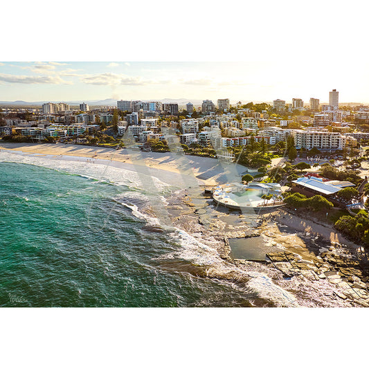 The AWP Image Librarys Caloundra Kings Beach Drone Image 0237 captures a coastal city with a sandy stretch, turquoise ocean pool, high-rise buildings lining the beach, and a unique rounded structure near the water, all under a clear sky and warm sun.