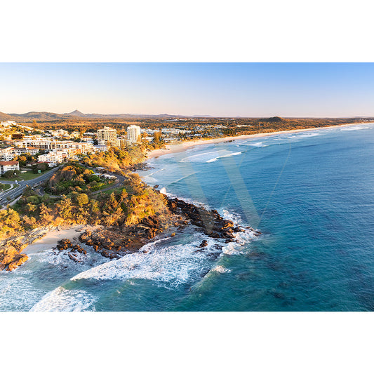 The Coolum Beach Drone Image 0647 by AWP Image Library captures an aerial view of a coastal town with buildings near a sandy beach, flanked by rocky cliffs and lush greenery. The clear sky extends over the ocean, where waves softly meet the shore.