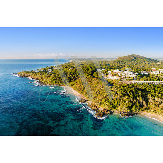 The Coolum Beach Drone Image 0932 from AWP Image Library captures a coastal landscape with winding shorelines, lush green hills, and a blue ocean. Buildings blend into the greenery under a clear, bright sky.