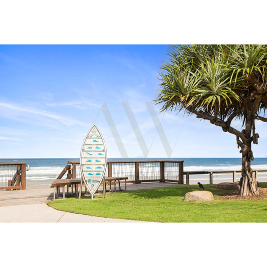 The Coolum Beach Image 6127 from the AWP Image Library showcases a stunning beach with a clear blue sky, sandy shore, and ocean waves. It features a surfboard-shaped sign with fish images, a tropical tree on green grass, and a wooden boardwalk nearby.