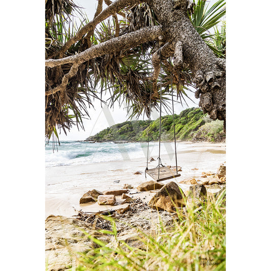 The Coolum Beach Image 6366 from AWP Image Library depicts a wooden swing hanging from a tree on a tropical beach, rocks and sand along the shoreline, waves crashing, and lush green foliage under a cloudy sky.