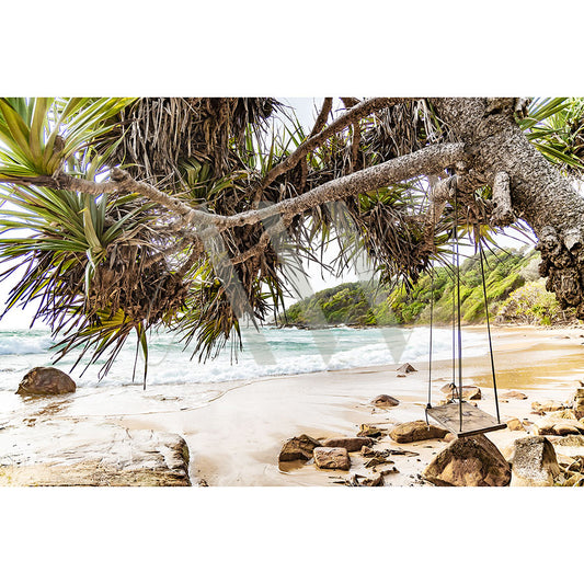 The Coolum Beach Image 6403 from AWP Image Library portrays a tranquil, tropical scene with a rustic tree swing on a gentle, sandy shoreline. Lush greenery and large rocks surround the area while sunlight filters through the leaves, enhancing the peaceful atmosphere.