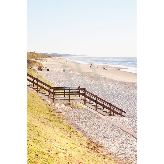 Coolum Beach Image 9262 by AWP Image Library captures First Bay on the Sunshine Coast, with a sandy beach, gentle waves, and a wooden pathway descending from a grassy area. People enjoy the sunny day on the shore and swings nearby under a clear sky with an ocean stretching to the horizon.