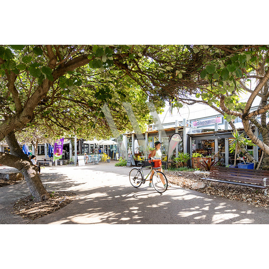 A person strolls with a bicycle in the shade of trees, surrounded by cafes and shops. Sunlight dapples the path, while vibrant signs and outdoor seating create a lively atmosphere akin to Coolum Beachs appeal. Captured in Coolum Beach Main St Image 6166 by AWP Image Library.