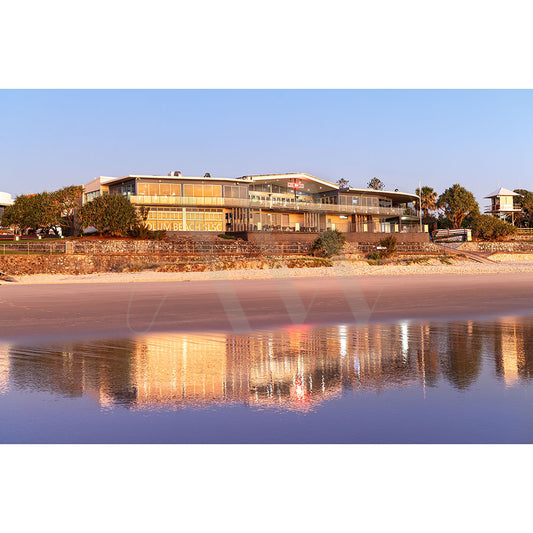 The Coolum Beach Surf Club Image 8763 from AWP Image Library features a modern building with large windows, a sign by the beach reflecting in calm sunset waters, surrounded by trees and sandy shores.