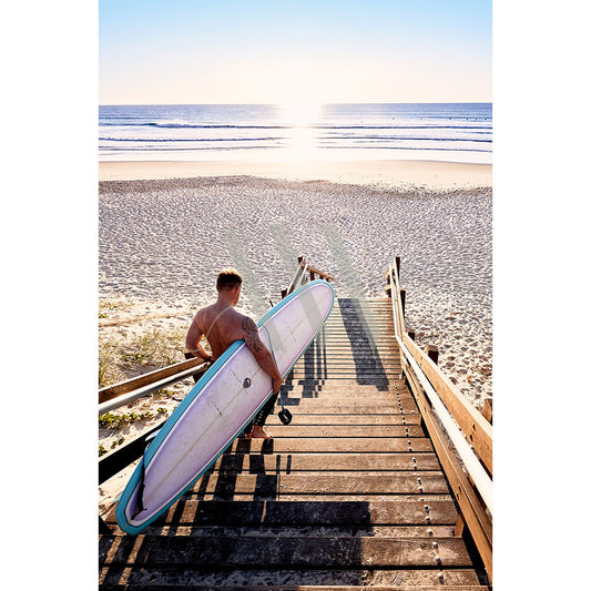 A person with a Coolum Beach Surfer 9103 surfboard from AWP Image Library descends wooden stairs to a sandy beach. The low sun casts a warm glow on the ocean waves, suggesting a serene early morning or late afternoon surf session.