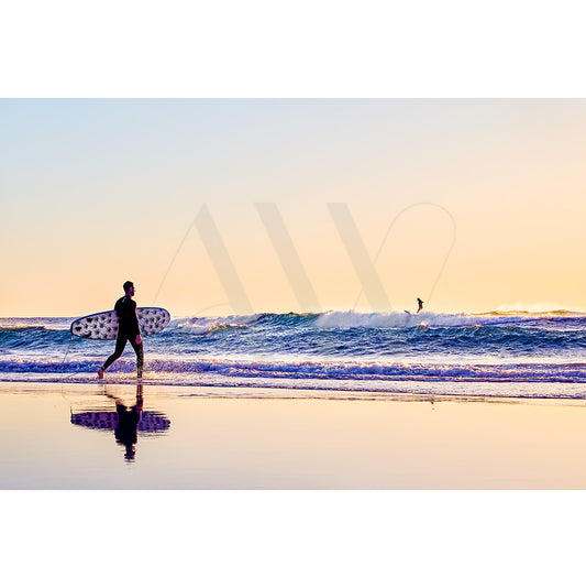 The Coolum Beach Surfers Image 9061 by AWP Image Library captures a surfer with a polka dot surfboard walking along the shoreline at sunrise, while another surfer rides a distant wave under a serene, golden sky.