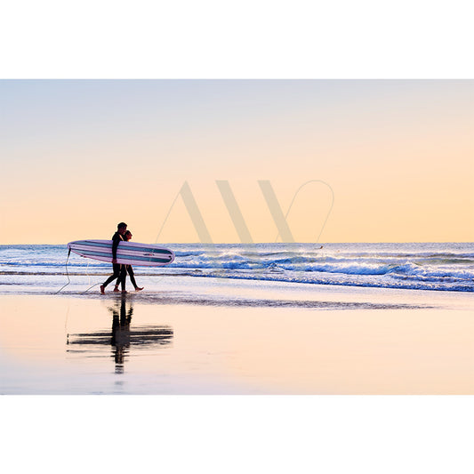 Two surfers wander along the shoreline, surfboards in hand while gentle waves graze their feet. The sky, a delicate blend of orange and blue at sunset, mirrors upon the damp sand. 