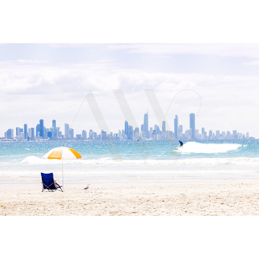 Currumbin Beach Image 4451 by AWP Image Library shows a sandy beach with a yellow and white umbrella, a blue chair, a nearby seagull, a surfer riding a wave in the background, and a city skyline under partly cloudy skies.