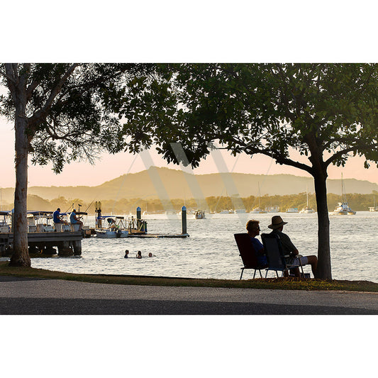 As the sun sets by the Noosa River, two people sit under a tree on Gympie Terrace, admiring swimmers and docked boats with softly lit hills in the distance. Capture this serene scene with Gympie Tce Image 7359 from AWP Image Library.