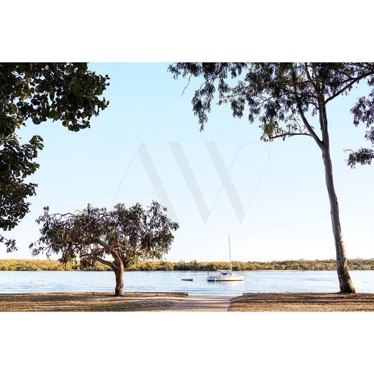 The Gympie Tce Image 7934 by AWP Image Library captures a serene lake with a sailboat anchored near the shore. Trees frame the scene, casting shadows on the grassy foreground, and a clear blue sky enhances its peacefulness.