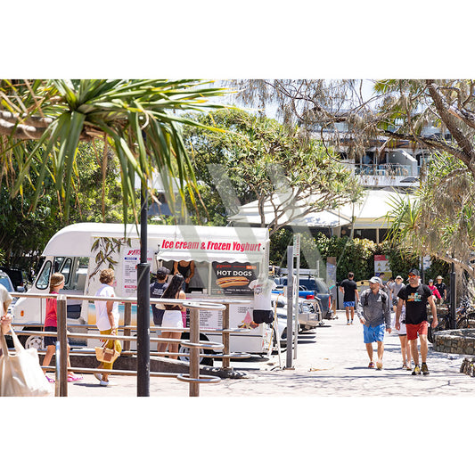 Hastings Street Image 4939 by AWP Image Library captures a vibrant outdoor scene near Noosas Main Beach with people passing an ice cream and frozen yogurt truck. Palm trees and greenery frame the area, with a hot dog sign nearby, all under sunny skies perfect for enjoying sweet treats.