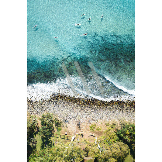 The Noosa Drone Image 0055 by AWP Image Library captures an aerial view of a shoreline with clear turquoise water, gentle waves, and swimmers. A pebble beach meets lush green trees with a small path and bench visible on the grass.