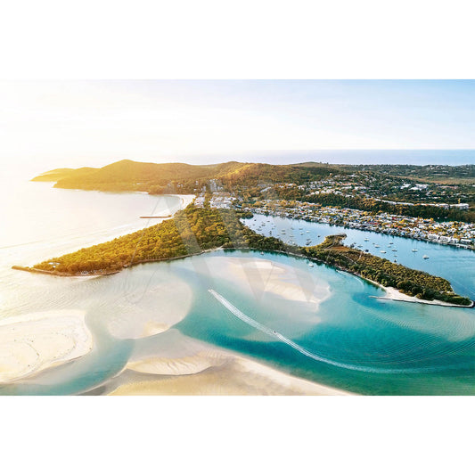 Experience the coastal charm captured in Noosa Drone Image 0095 by AWP Image Library, featuring an aerial view of the Noosa River winding toward the ocean. The image showcases sandy shores, lush greenery, and boats docked near a picturesque small town, all illuminated by sunlight that brightens the horizon, creating a serene atmosphere.