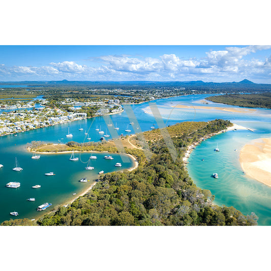 Noosa Drone Image 0811 by AWP Image Library captures an aerial view of sailboats on turquoise and dark blue waters, sandy beaches, and lush green vegetation. A town lies in the background beneath a partly cloudy sky.