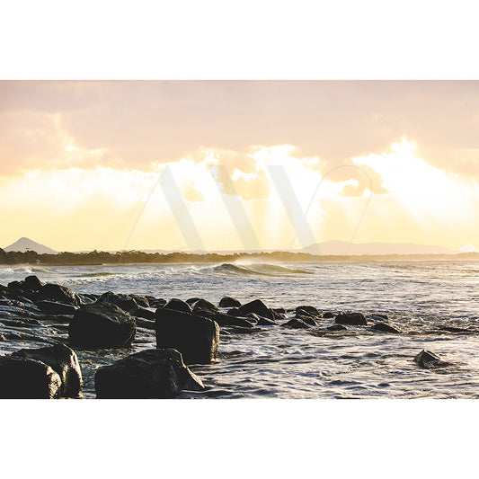 Noosa Image 3221 from AWP Image Library captures a serene coastal scene at sunset, with waves softly crashing onto a rocky beach. The sky glows in warm orange and yellow tones as the sun peeks through low clouds, offering a picturesque view.