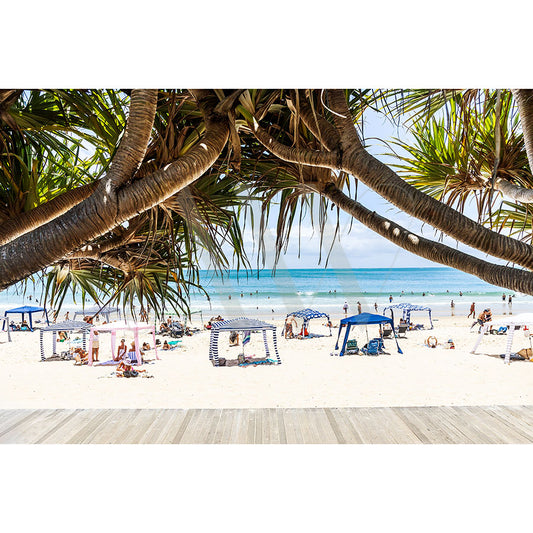 The Noosa Main Beach Image 417 from AWP Image Library captures a sunny beach scene with people under blue and white tents on the sand, a visible ocean background with gentle waves, swimmers in the water, and palm tree branches framing the top of the image.