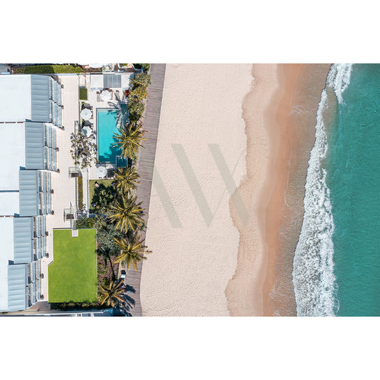 The Noosa Main Beach Drone Image 116 by AWP Image Library captures an aerial view of luxury beachfront resorts with modern white buildings, a swimming pool, and palm trees on the left, set against the sandy shores and turquoise ocean waves of Noosa Beach on the right.