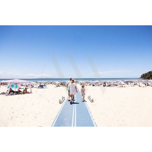 A family strolls along a boardwalk toward Noosa Main Beach (Image 4771) by AWP Image Library, with tents and umbrellas dotting the sand under a clear blue sky. The ocean and a tree-covered hill are visible in the background.