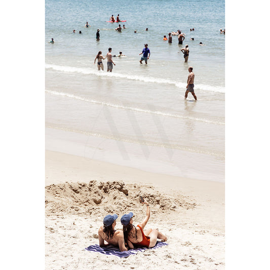 Two people sit on a blanket at Noosa Main Beach, wearing hats and swimsuits, gazing at the ocean. In the background of AWP Image Librarys Image 4853, several people swim and stand in shallow water under a bright sky.