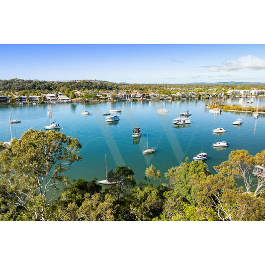 The Noosa River Drone Image 0273 by AWP Image Library features a tranquil coastal scene with calm blue water, docked boats, lush green trees in the foreground, and houses along the distant shore under a clear sky.