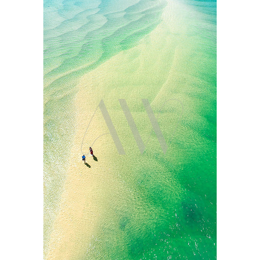 Product: Noosa River Drone Image 0856 by AWP Image Library shows an aerial view of two people walking on a sandbank, casting long shadows in the sunlight against the clear turquoise water, creating a striking contrast with the sandy path.