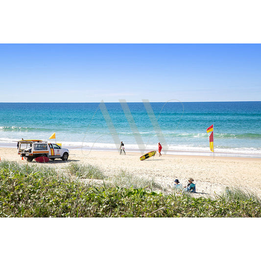 The Sunshine Beach Image 2502 by AWP Image Library captures a sunny beach scene with a lifeguard truck on the sand, two flags marking a safe swimming zone, two people walking, one holding a surfboard, and two individuals sitting on grass overlooking the beach.