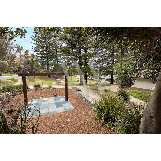 The serene outdoor garden at Sunshine Beach Playground (Image 2522) from AWP Image Library features a checkered patio, mulch, and various plants, ideal for leisure activities. Tall trees and lush greenery offer shade with a pathway and benches in the background.