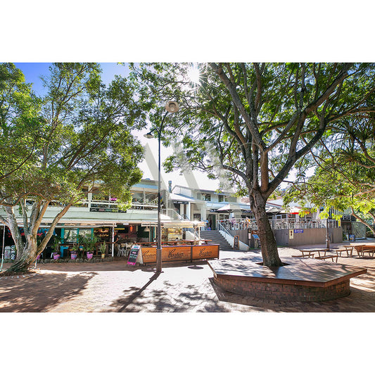 Sunshine Beach Shops Image 2503 by AWP Image Library depicts a sunny plaza with trees, benches, and a brick walkway. The scene includes a cafe with outdoor seating and various small shops in a tranquil two-story building shaded by trees.